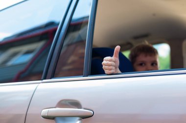Çocuk gümüş renkli bir arabanın arka koltuğunda oturur ve başparmağını gösterir. Anaokulu öğrencisi heyecanlı bir geziye hazır..