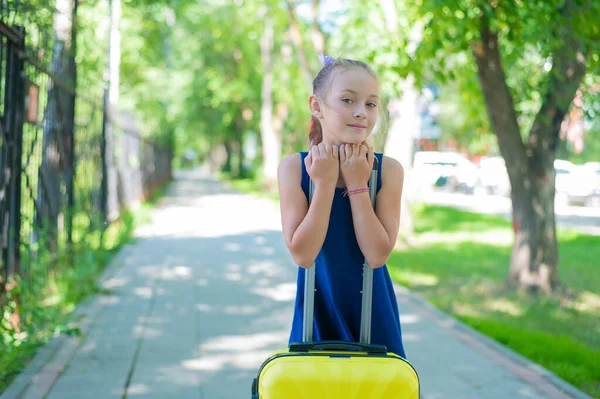 Een tevreden meisje in een blauwe jurk staat alleen op straat met een gele koffer en wacht op haar ouders. Een kind gaat op zomervakantie.. — Stockfoto