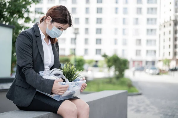 Depressed woman in medical mask sits on parapet outdoors with a box of personal items from the desktop. Unemployed girl alone on the street. Bankruptcy of a company due to quarantine in coronavirus.