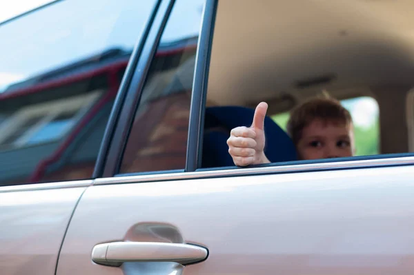 Çocuk gümüş renkli bir arabanın arka koltuğunda oturur ve başparmağını gösterir. Anaokulu öğrencisi heyecanlı bir geziye hazır.. — Stok fotoğraf