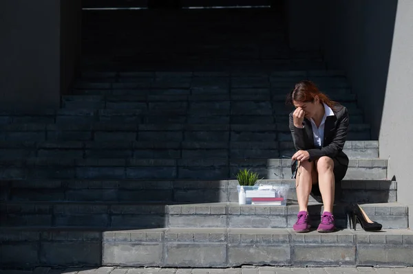 Mujer deprimida vestida con máscara médica es despedida y está sentada en las escaleras con una caja de pertenencias personales. Trabajadora de oficina con traje y zapatillas al aire libre. El desempleo en la crisis económica —  Fotos de Stock