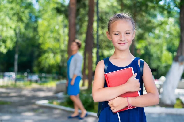 Das Porträt einer fleißigen Schülerin mit Rucksack und Buch geht in die erste Klasse. Mutter schickt glückliche Tochter in die Grundschule. — Stockfoto