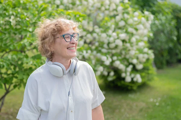 Une femme caucasienne âgée marche dans un parc et écoute de la musique. Une pensionnée souriante enlève son casque et profite de l'arôme des arbres à fleurs dans le jardin. — Photo