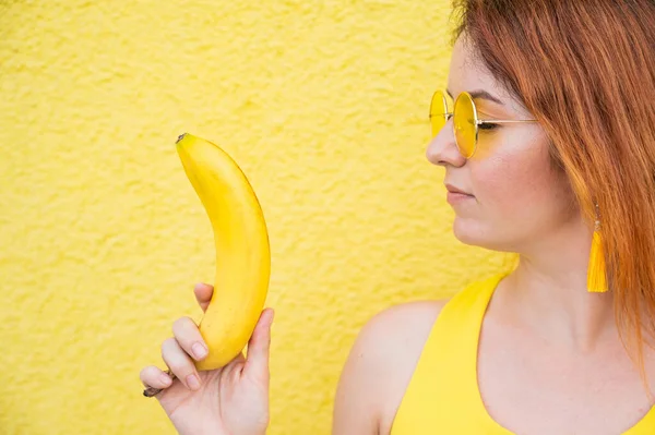 손에 바나나를 들고 선글라스를 쓴 백인 여성의 모습. 노란 배경에 잘 어울리는 매력적 인 아가씨. — 스톡 사진