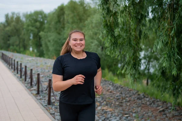 Een zwaarlijvige jonge vrouw die buiten jogt. Fat mooi lachend meisje in een zwart trainingspak is bezig met fitness voor gewichtsverlies aan het water. Een vrouw rent op een zomerdag. — Stockfoto