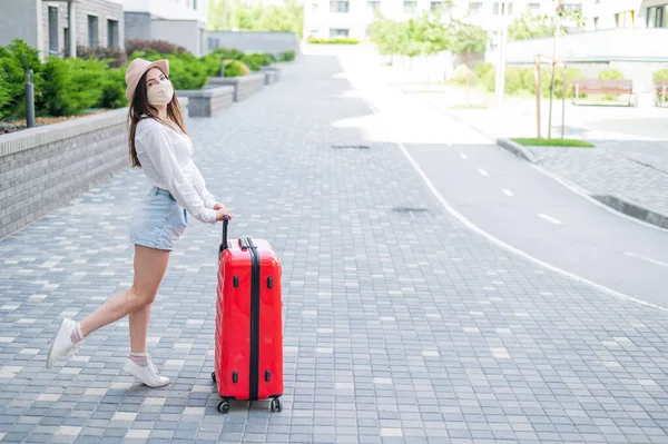 Красивая молодая белая женщина в медицинской маске путешествует с красным чемоданом по пустому городу. Девушка отправляется в отпуск с большой сумкой во время эпидемии коронавируса. — стоковое фото