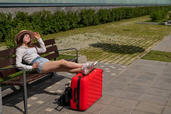Die schöne kaukasische Frau mit Hut und kurzen Hosen sitzt auf einer Bank und streckt ihre Beine auf einem roten Koffer aus und wartet auf ein Taxi. Mädchen wartet auf Abreise auf Sommerreise. — Stockfoto