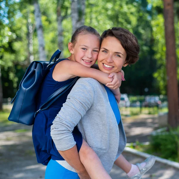 Una bella donna caucasica si alza all'aperto e tiene la figlia sulla schiena. Una allegra ragazza sorridente siede sulla schiena della madre mentre cammina nel parco. Famiglia amorevole amichevole. — Foto Stock