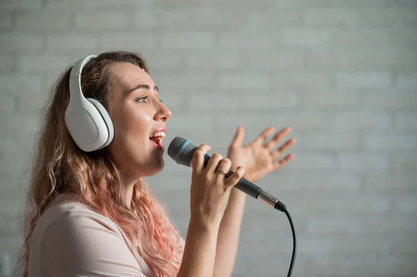 Közelkép egy göndör hajú kaukázusi nőről, aki mikrofonba énekel. Gyönyörű érzelmi lány fehér fülhallgatóval énekel egy dalt otthon karaoke és aktívan gesztusok ellen téglafal. — Stock Fotó