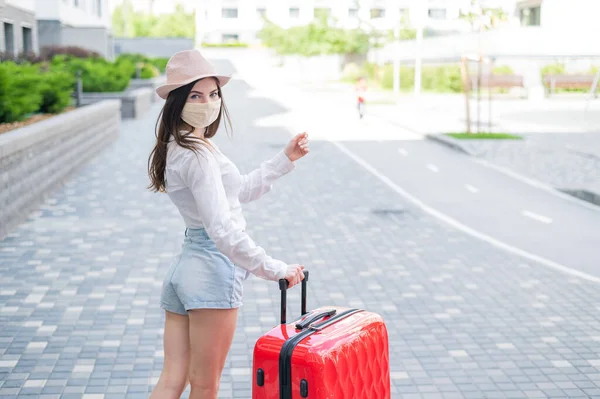 Een mooie jonge blanke vrouw met een medisch masker reist met een rode koffer door een lege stad. Het meisje gaat op vakantie met een grote bagage tas tijdens de coronavirus epidemie. — Stockfoto