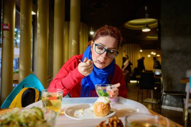 Kafede salata yiyen kızıl saçlı beyaz bir kadın. Gözlüklü ve mavi eşarplı düşünceli romantik kız yemek yiyor..