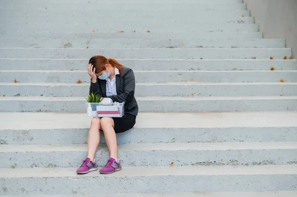 Una mujer con una caja de pertenencias personales se sienta en las escaleras y dobla las manos desesperada en la cabeza. Empleada con máscara médica despedida debido a crisis financieras en coronovirus. Desempleo —  Fotos de Stock