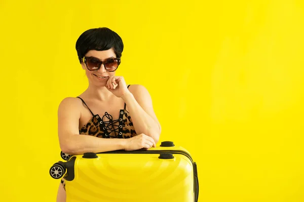 Портрет привлекательной женщины в цельном купальнике леопарда и солнечных очках с чемоданом на желтом фоне. Соблазнительная брюнетка в монокини готова отправиться в море. Летний отдых. — стоковое фото