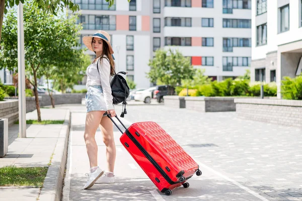 Schöne junge kaukasische Frau mit Sommermütze posiert mit einem roten Koffer im Freien. Glücklich lächelndes Mädchen geht mit einer großen Gepäcktasche in den Urlaub. Charmante Dame in kurzen Hosen an einem heißen, sonnigen Tag. — Stockfoto