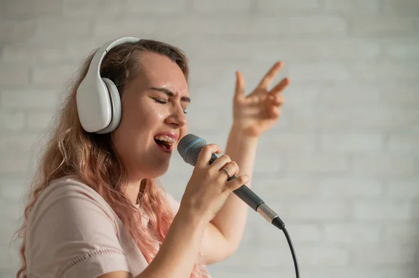 Közelkép egy göndör hajú kaukázusi nőről, aki mikrofonba énekel. Gyönyörű érzelmi lány fehér fülhallgatóval énekel egy dalt otthon karaoke és aktívan gesztusok ellen téglafal. — Stock Fotó