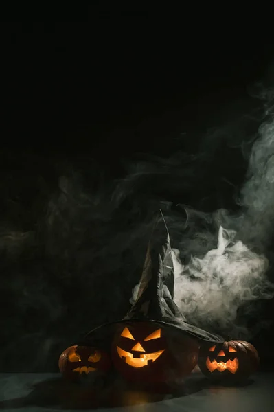 Carte verticale Halloween. Chapeau de sorcière sur une citrouille avec grimaces effrayantes sculptées sur un fond noir dans le brouillard. Jack-o-lanterne brille de l'intérieur et émet de la fumée. — Photo