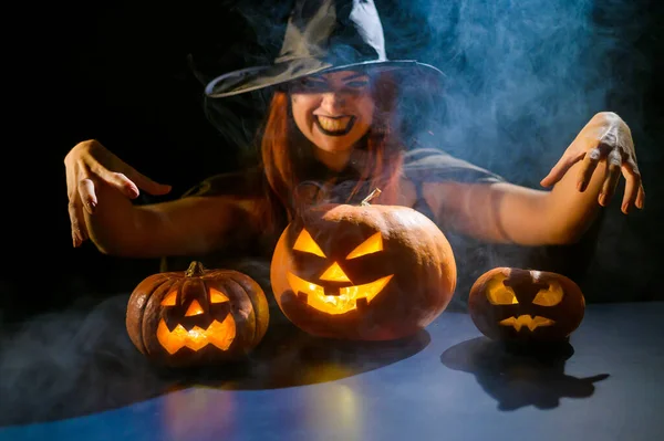 Złowroga wiedźma w kapeluszu przywołuje latarnię. Tradycyjne postacie halloween. Mistyczna mgła skrada się nad dyniami z wyrzeźbionymi okropnymi twarzami. Kobieta w stroju karnawału. — Zdjęcie stockowe