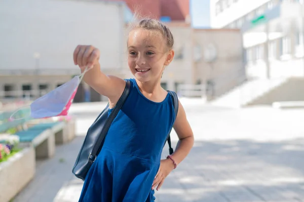 A colegial feliz tira a máscara. Menina sorridente alegre com uma mochila remove a máscara e vai para a escola. Fim da quarentena e retorno à vida normal . — Fotografia de Stock