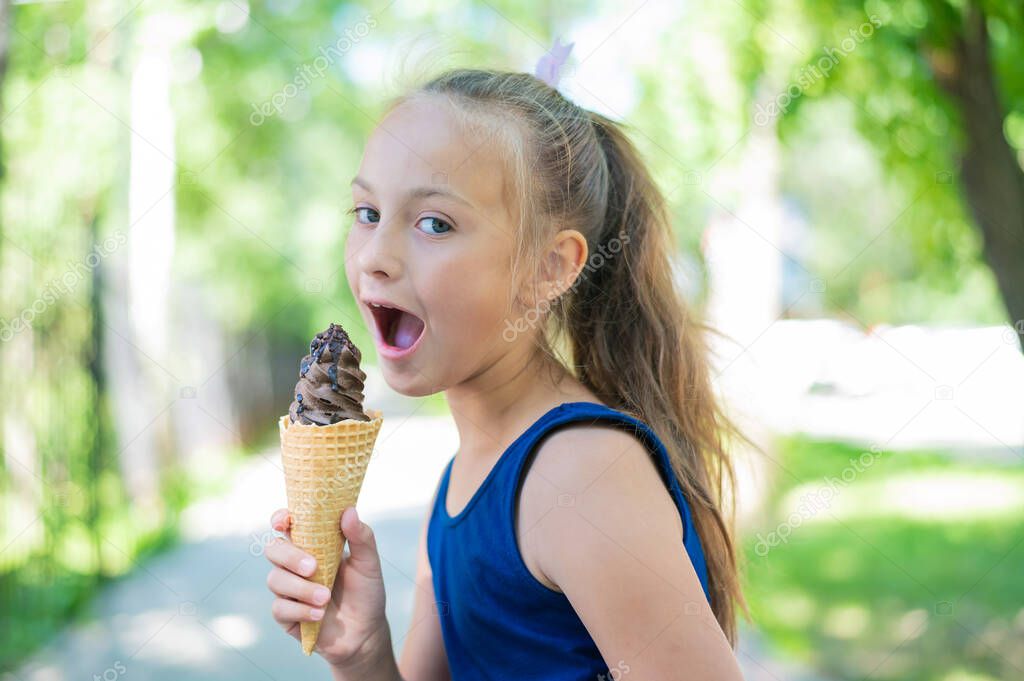 Happy little caucasian girl in blue dress eats chocolate ice cream cone outdoors. An emotional excited child enjoys a cooling gelato on a hot summer day.