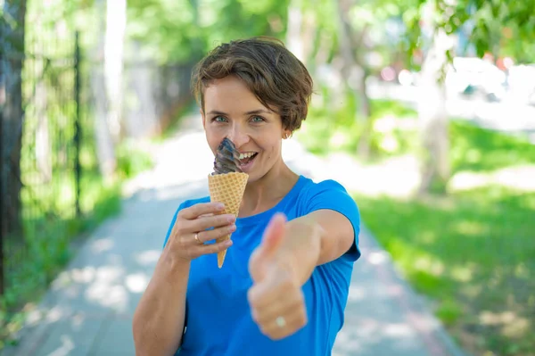 Очаровательная белая женщина с короткой стрижкой улыбается, ест мороженое и показывает большой палец. Эмоциональная девушка с красивой улыбкой наслаждается мороженое прогулки в парке. — стоковое фото