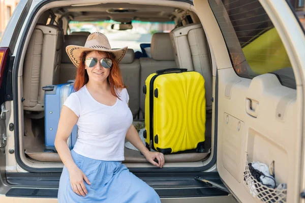 Glückliche rothaarige Frau mit Cowboyhut und Sonnenbrille auf einer Autofahrt durch das Land. Mädchen im Kofferraum mit gelbem Koffer, bereit für den Sommerurlaub. Unabhängiges Reisen. — Stockfoto