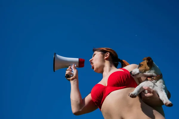 Кавказька жінка в червоному бікіні працює рятувальником на пляжі і кричить через мегафон проти блакитного неба. Дівчинка тримає цуценя-джека в русселі тер'єра і гучномовця. Літня відпустка. — стокове фото