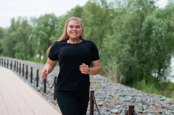 Eine übergewichtige junge Frau joggt im Freien. Dicke schöne lächelnde Mädchen in einem schwarzen Trainingsanzug ist in Fitness für die Gewichtsabnahme am Wasser engagiert. Eine Frau läuft an einem Sommertag. — Stockfoto