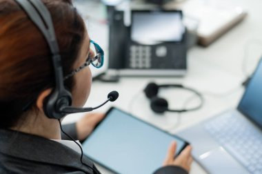 Masada otururken dijital tablet kullanan kulaklıklı bir kadının arka görüntüsü. İş yerinde dost canlısı kadın destek operatörü.