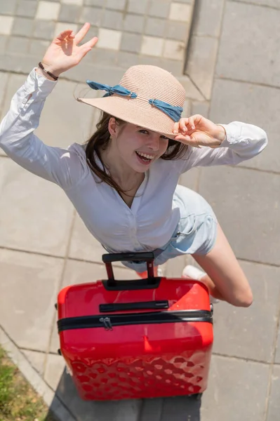 Top uitzicht op mooie jonge Kaukasische vrouw in de zomer hoed poseren met rode koffer buiten. Gelukkig lachend meisje gaat op vakantie met een grote bagage tas. Charmante dame in korte broek op een hete zonnige dag. — Stockfoto