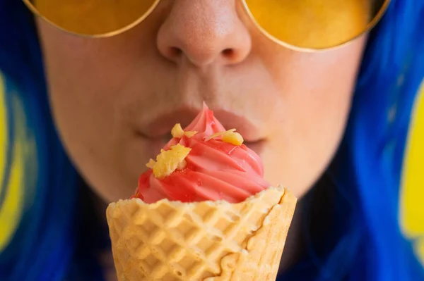 Closeup of sensual female lips with a waffle cone of pink ice cream. Cropped photo of a woman in sunglasses with blue hair happily eating pink gelato on a hot summer day. Macro. Happy sweet tooth.