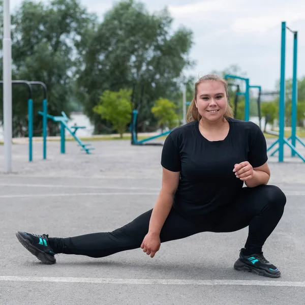 Fettleibige junge Kaukasierin beim Seitensprung. Dicke schöne lächelnde Mädchen in einem schwarzen Trainingsanzug ist in Fitness im Freien beschäftigt. Aufwärmen vor dem Training. — Stockfoto