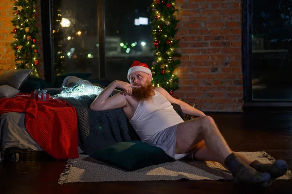Image humoristique d'un homme sans pantalon devant Noël. Le Père Noël chauve avec une barbe rouge déballe un cadeau. Une parodie d'une séance photo de Nouvel An pour femmes glamour. Blague. — Photo