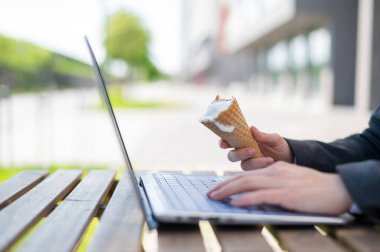 Yüzü olmayan bir kadın yazlık terasta bilgisayar başında çalışıyor ve vanilyalı dondurma yiyor. Dizüstü bilgisayarda yazı yazan ve elinde dondurma külahı tutan kadınların yakın çekimi. Yaz serinletici tatlı.