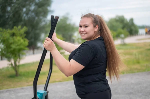 Schöne lächelnde übergewichtige junge Frau macht Fitness auf einem Ellipsoid-Simulator im Freien. Dicke Mädchen trainieren auf dem Sportplatz zum Abnehmen. Sport draußen an einem warmen Sommertag. — Stockfoto