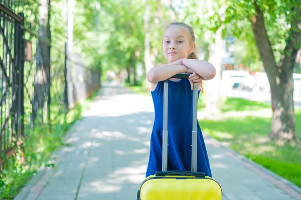 Een tevreden meisje in een blauwe jurk staat alleen op straat met een gele koffer en wacht op haar ouders. Een kind gaat op zomervakantie.. — Stockfoto