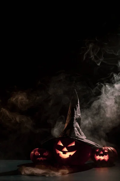 Biglietto di Halloween verticale. Cappello da strega su una zucca con smorfie raccapriccianti scolpite su fondo nero nella nebbia. Jack-o-lanterna brilla dall'interno ed emette fumo. — Foto Stock