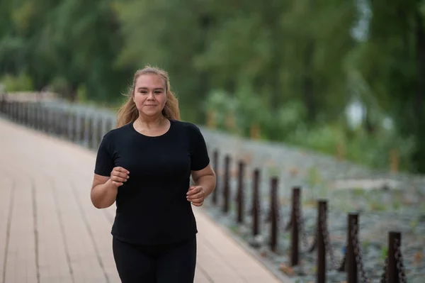 Una joven obesa corriendo al aire libre. Gorda hermosa chica sonriente en un chándal negro se dedica a la aptitud para la pérdida de peso en el paseo marítimo. Una mujer corre en un día de verano. — Foto de Stock