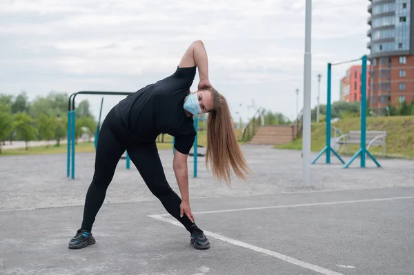 Une jeune femme en surpoids portant un masque de protection s'entraîne sur le terrain de sport. Une fille obèse fait des pentes à l'extérieur. Faire du sport en quarantaine. — Photo