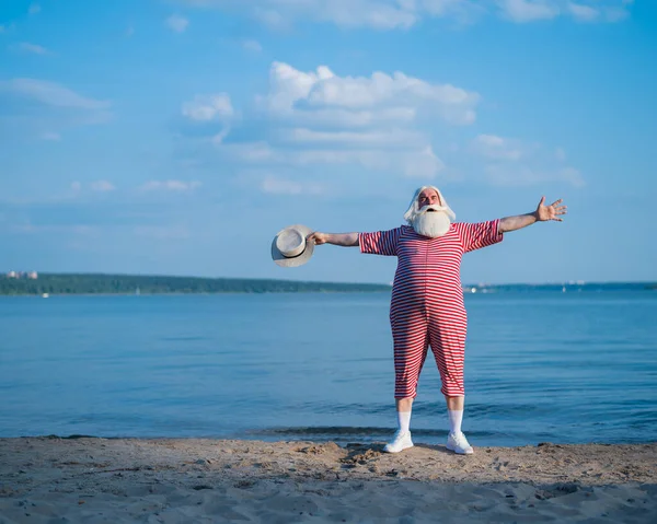 Пожилой седой мужчина с бородой в полосатом купальнике и шляпе, позирующий на пляже. Пенсионер на отдыхе у озера. — стоковое фото