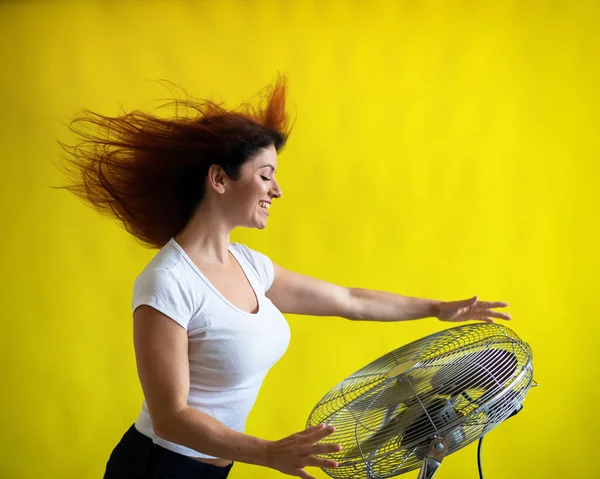 Una hermosa mujer pelirroja se enfría de pie sobre un gran ventilador eléctrico sobre un fondo amarillo. Chica con el pelo en desarrollo en el viento. Dispositivo para enfriar el aire. — Foto de Stock