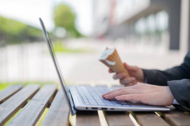 Yazlık terasta dizüstü bilgisayarlı bir klavyeyle bir iş kadınının ellerine yakın çekim. Ofis görevlisi dışarıda dondurma külahı yiyor. Kesilmiş fotoğraf. Vanilyalı dondurulmuş yoğurt..