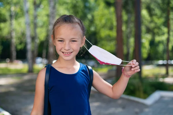 Bonne petite fille enlève le masque à l'extérieur. Joyeuse écolière souriante avec sac à dos enlève le masque et va à l'école. Fin de la quarantaine et retour à la normale. — Photo