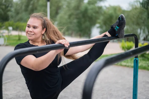 Grosse fille est engagée dans l'étirement sur les barres inégales sur l'aire de jeux. Belle jeune femme souriante en surpoids faisant du sport pour perdre du poids à l'extérieur. — Photo