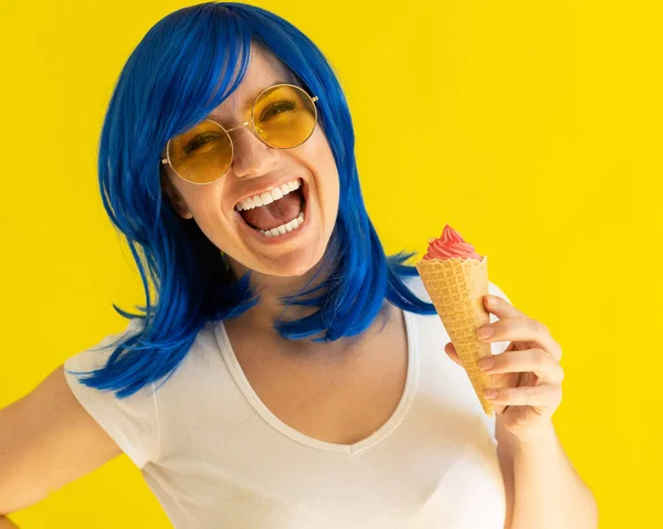 Портрет женщины с голубыми волосами в солнечных очках, держащей вафельный конус с мороженым на желтом фоне. Красивая кавказская девушка с прекрасной улыбкой наслаждается летним охлаждающим десертом. — стоковое фото