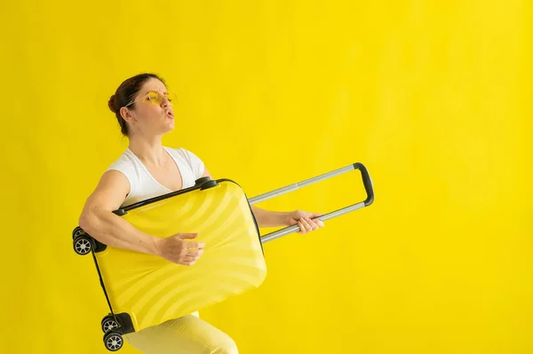 Smiling woman in sunglasses fooling around and holding a suitcase like a guitar on a yellow background. An excited girl in anticipation of a summer vacation trip simulates playing a string instrument. — Stock Photo, Image