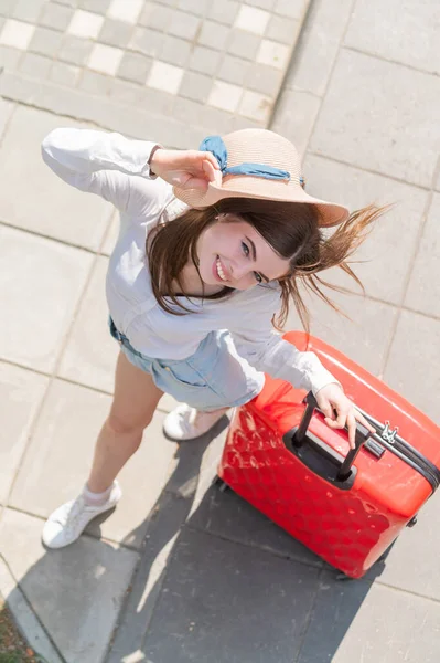 Top uitzicht op mooie jonge Kaukasische vrouw in de zomer hoed poseren met rode koffer buiten. Gelukkig lachend meisje gaat op vakantie met een grote bagage tas. Charmante dame in korte broek op een hete zonnige dag. — Stockfoto