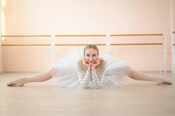 Όμορφη μπαλαρίνα στο σώμα και λευκό tutu είναι η κατάρτιση σε ένα μάθημα χορού. Νέοι ευέλικτοι χορευτές ποζάρουν σε pointe παπούτσια που κάθονται στο πάτωμα. — Φωτογραφία Αρχείου