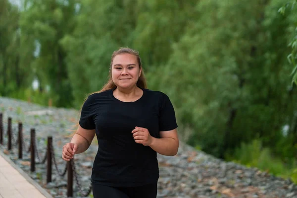En fet ung kvinna som joggar utomhus. Fett vacker leende flicka i en svart träningsoverall är engagerad i fitness för viktminskning vid vattnet. En kvinna springer på en sommardag. — Stockfoto