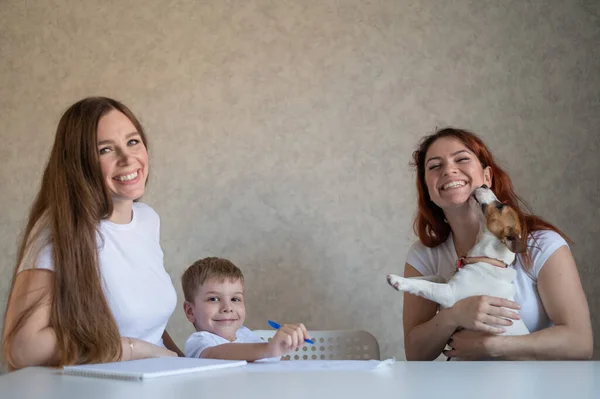 Happy family stays at home. Two women help the boy do school homework. Lesbian couple sitting at the table with their son and a cheerful puppy. Same-sex marriage with a child. Distance learning.