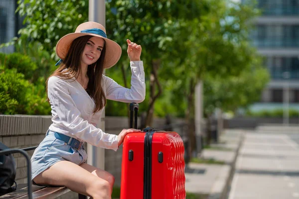 Eine schöne kaukasische Frau mit Hut und kurzen Hosen sitzt auf einer Bank und hält einen roten Koffer in der Hand und wartet auf ein Taxi oder einen Bus. Mädchen wartet auf Abreise auf Sommerreise. — Stockfoto